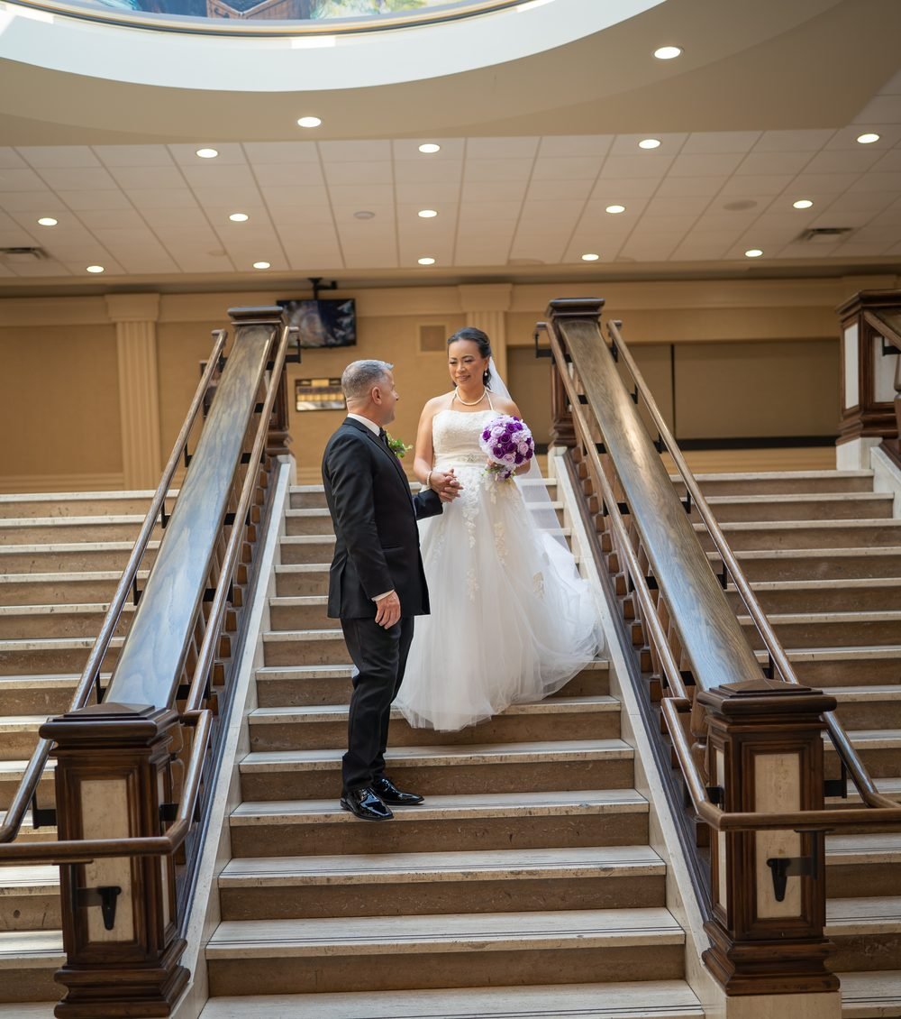 Groom taking his bride's hand on the grand staircase beneath the Caboto Club skylight